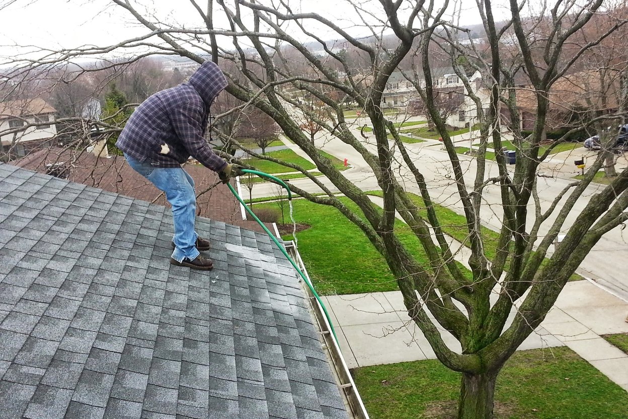 A solo contractor in work clothes photographing a finished gutter cleaning on a suburban house, with fallen leaves in the yard.